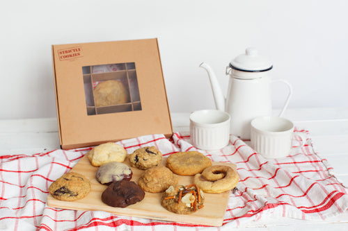 cookie box with a mix of cookies on a cutting board with 2 mugs and a pot of coffee